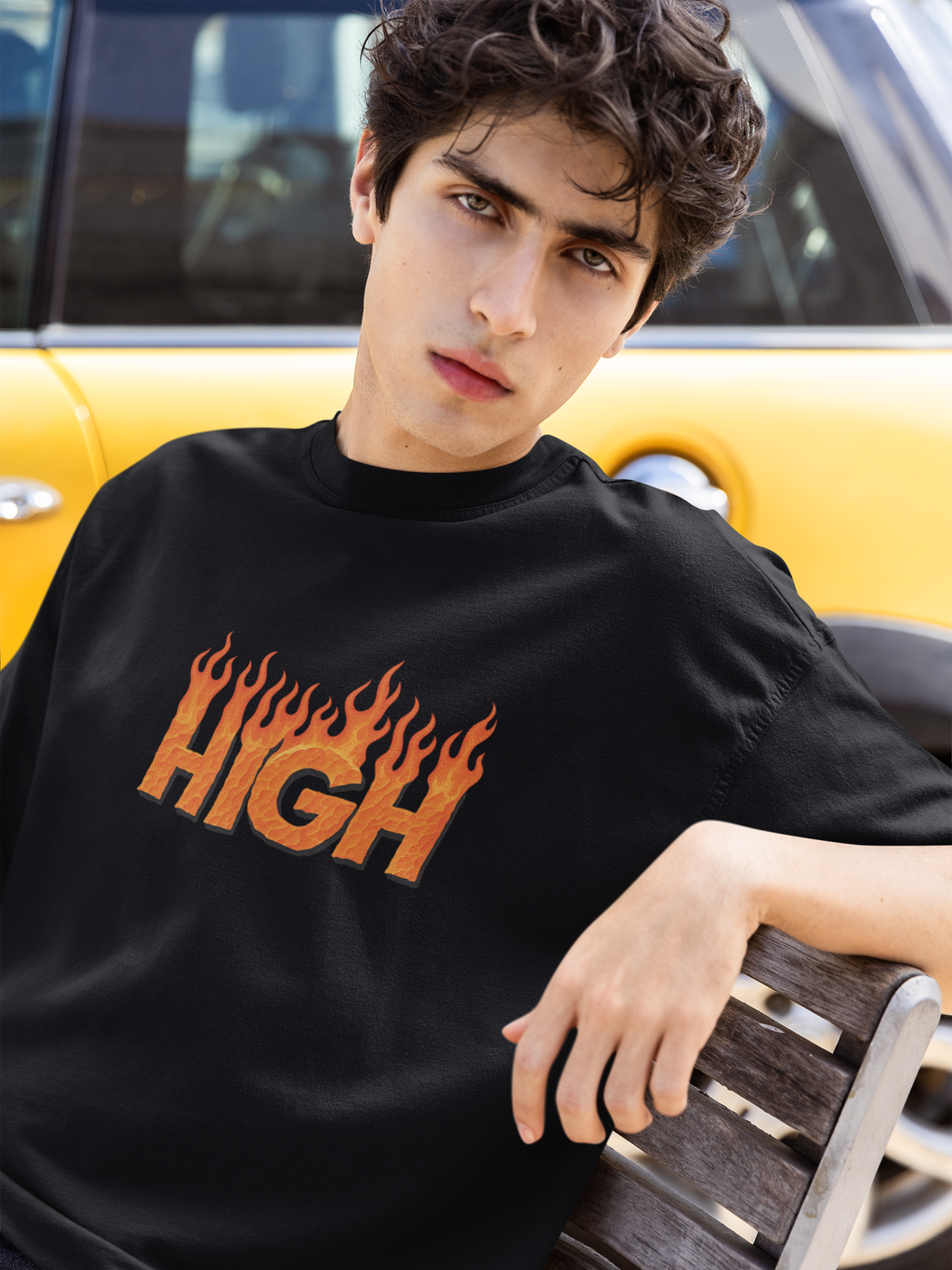Man wearing a black 240 GSM oversized t-shirt with flame-text "HIGH" graphic, sitting on a bench in front of a yellow car.