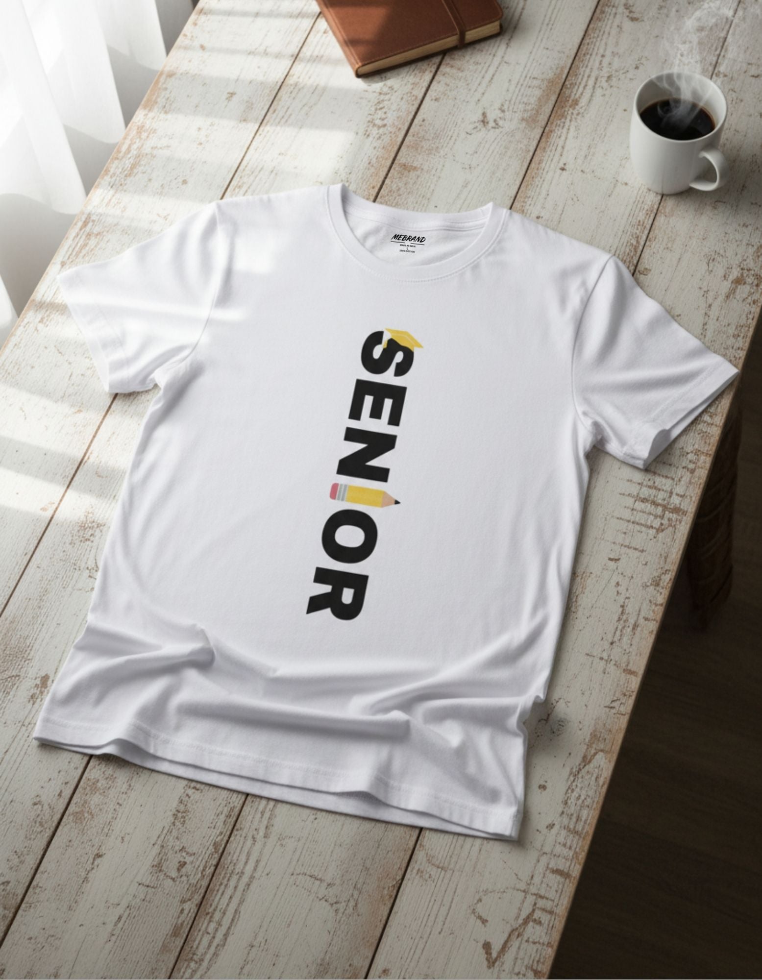 Young man wearing a white cotton T-shirt with "SENIOR" printed vertically in bold black letters, featuring a yellow graduation cap on the "S" and a colorful pencil design on the "I". Styled with light blue jeans for a casual look.