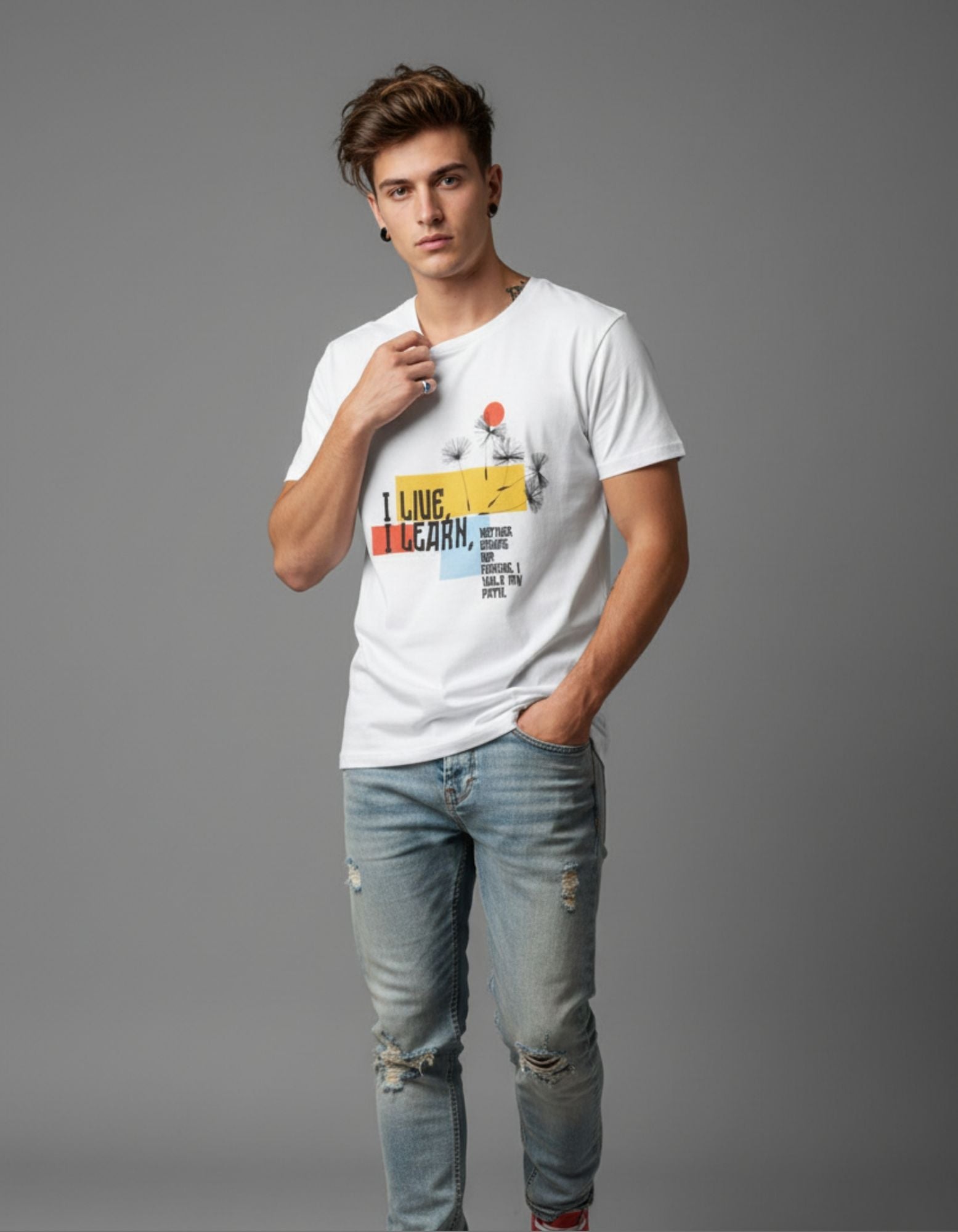Young man wearing a white graphic T-shirt with a bold quote design that reads “I Live, I Learn” in colorful typography, paired with light blue ripped jeans and red high-top sneakers. Styled in a casual, modern look against a plain grey background.