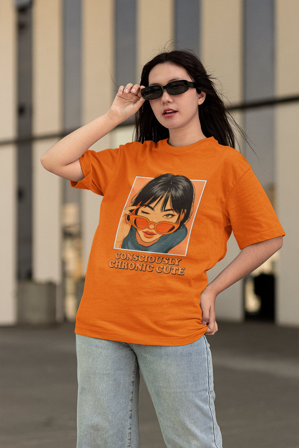 Woman wearing an oversized orange graphic t-shirt featuring an anime-style girl with sunglasses and the text "Consciously Chronic Cute", posing confidently with jeans and dark sunglasses.